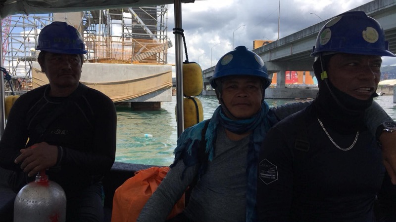 Three Oceantech divers on a boat in blue helmets and wetsuits with bridge construction behind them